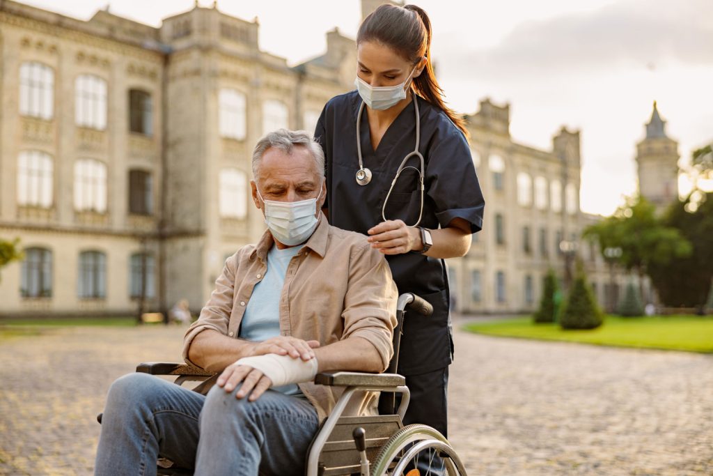 A nurse with a stethoscope assisting a masked senior man in a wheelchair outdoors.