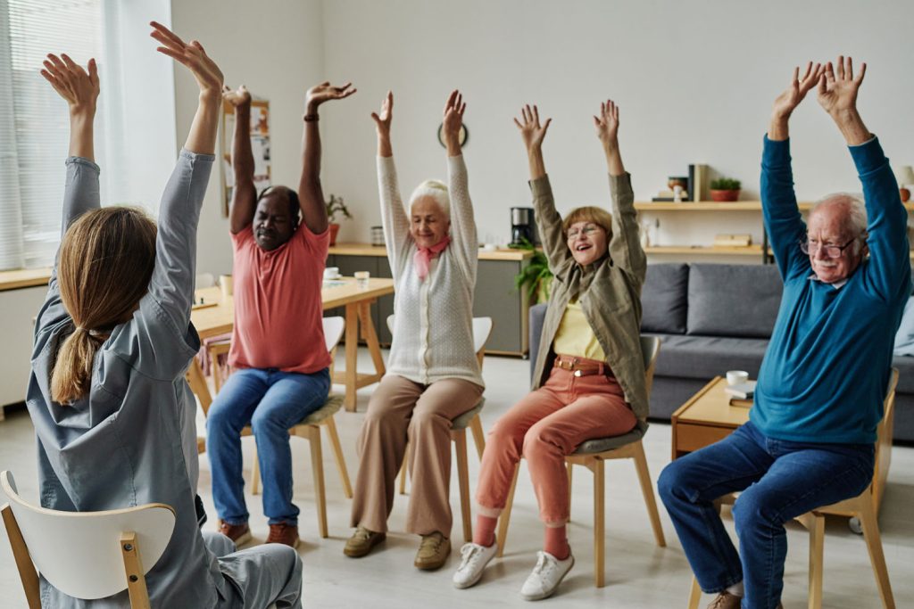 A group of aged people doing exercise sitting on wooden chair.